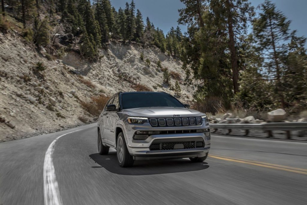 SUV Jeep Compass gris roulant sur une route sinueuse de montagne, entourée par des forêts de conifères sous un ciel clair.