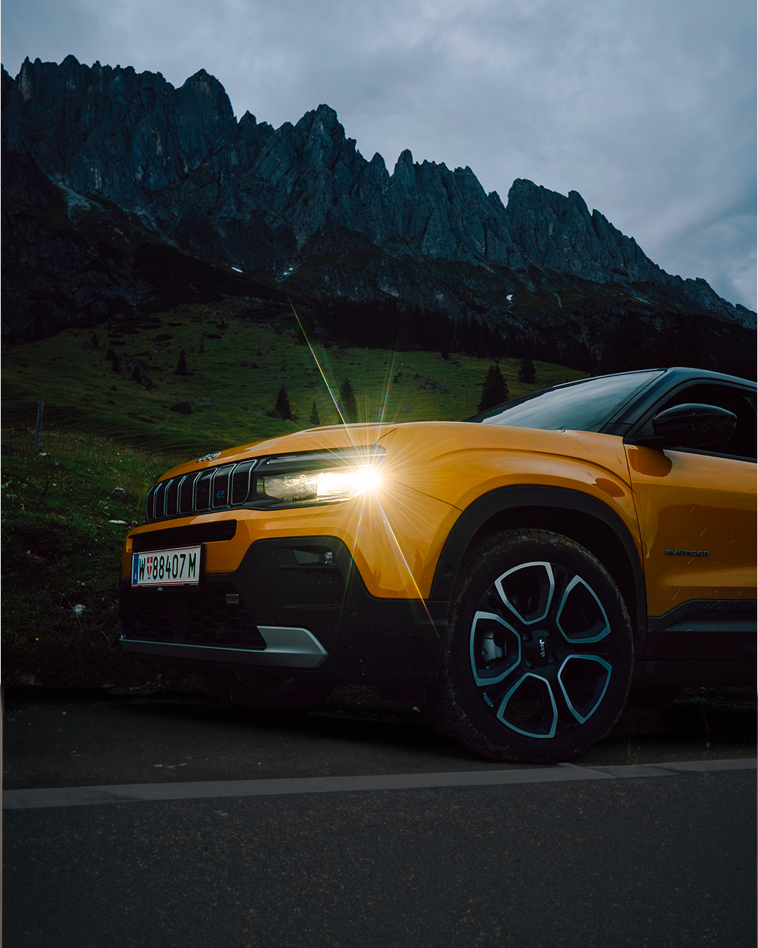 SUV Jeep Avenger jaune stationnée devant une chaîne de montagnes au crépuscule, phares allumés