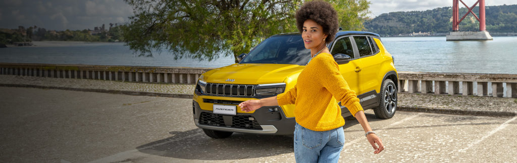 Femme marchant près d'une Jeep Avenger jaune garée près d'un pont et d'un plan d'eau